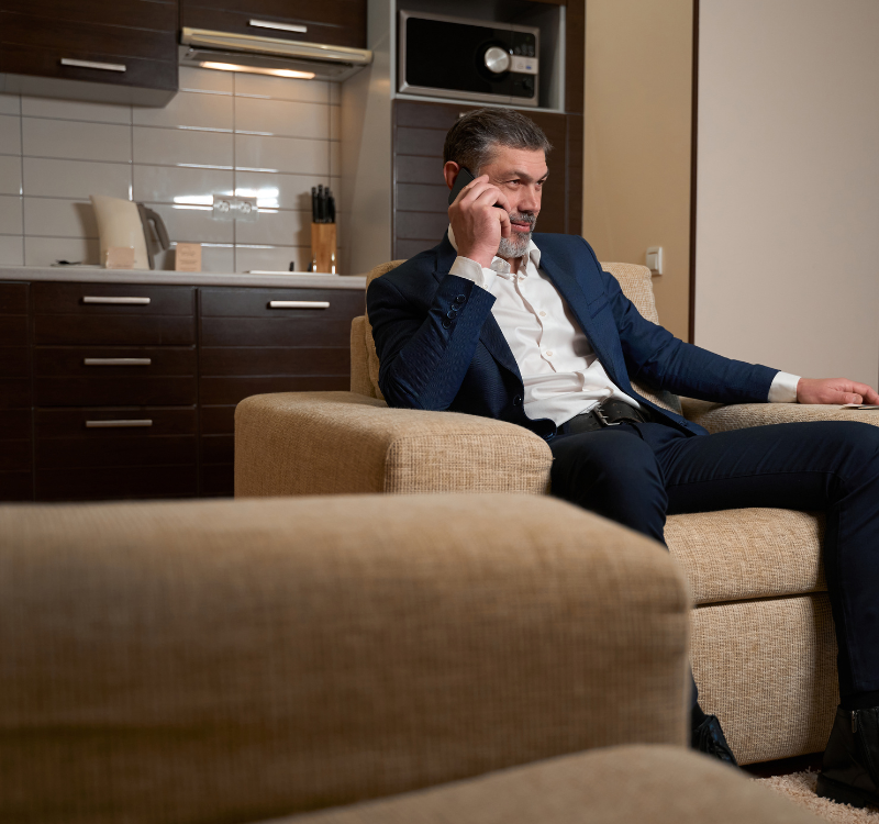 Corporate Business Housing The Ultimate Solution for Professionals on the Go (3) Businessman in a suit talking on the phone while seated in a modern furnished apartment with a full kitchen—illustrating Corporate Business Housing comfort.